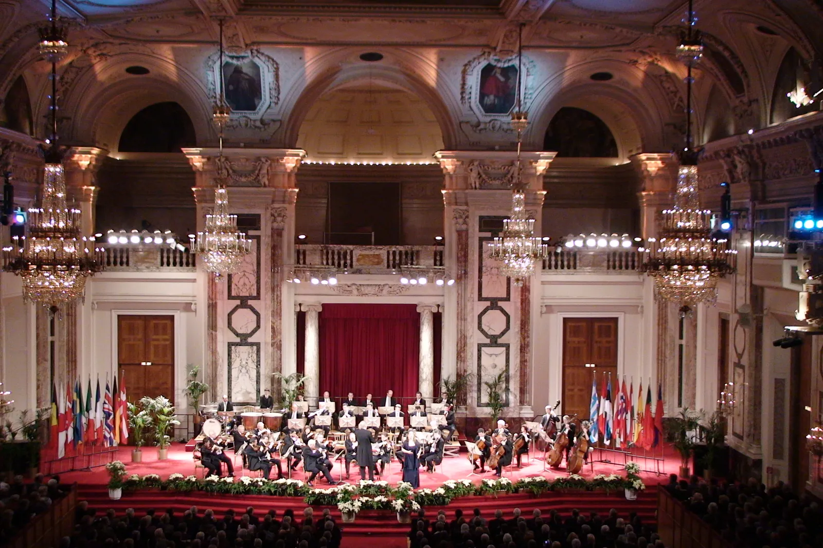The Hofburg Festival Hall from the gallery, with crystal chandeliers, red marble pilasters, a crimson curtain and the orchestra on a red-carpeted stage